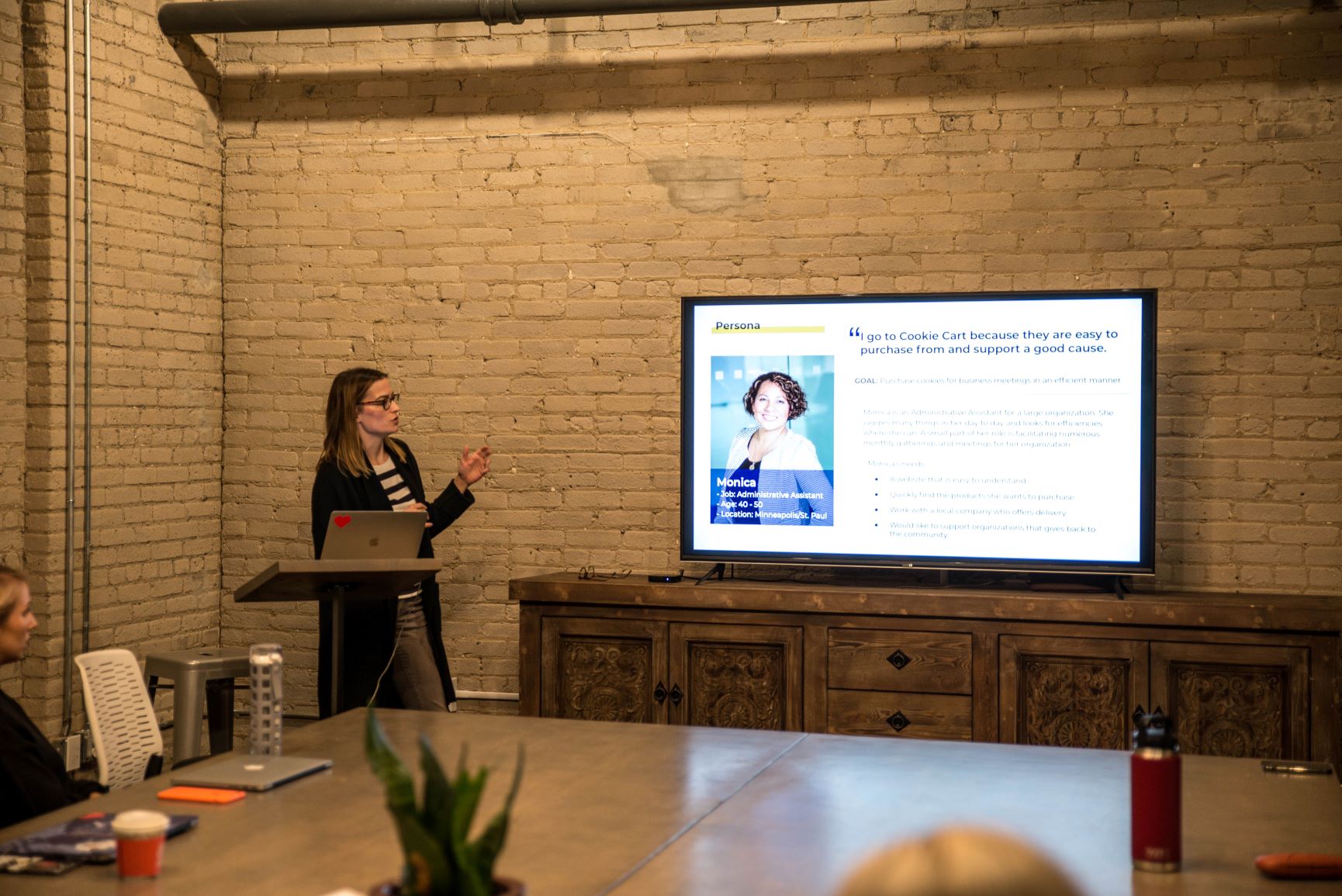 Woman standing behind a lectern facing a large screen with a persona projected on the screen